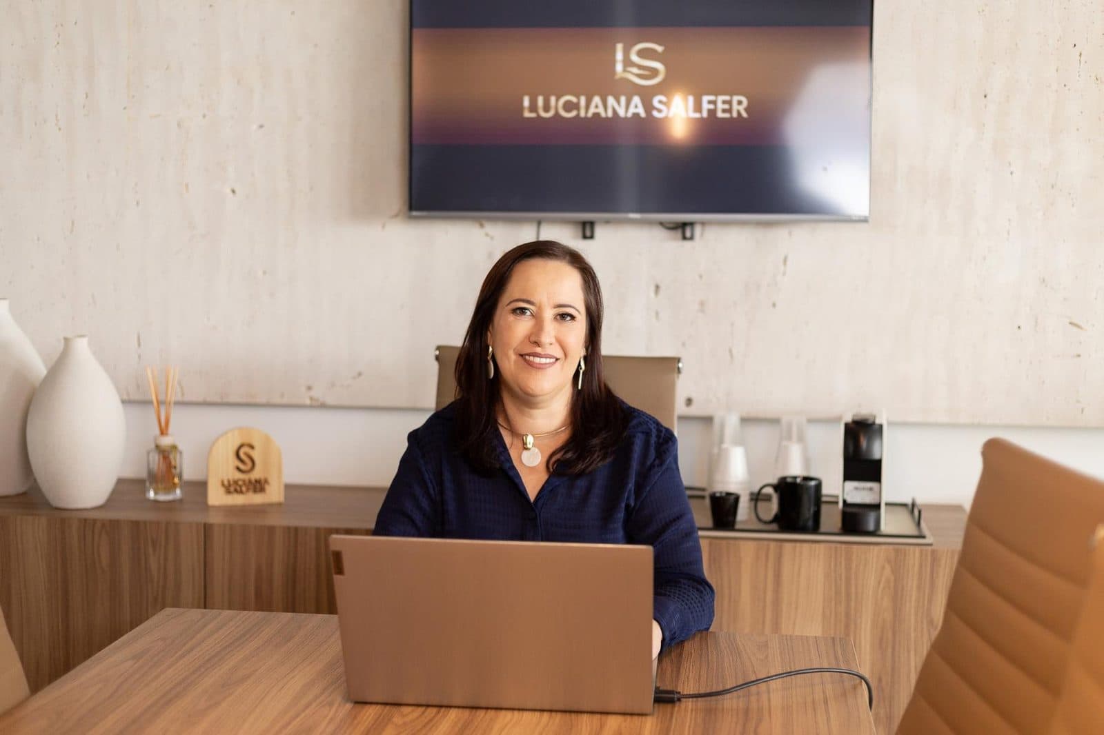 Luciana Salfer sentada à mesa com o notebook, sorrindo, com o logo da LS na TV atrás.