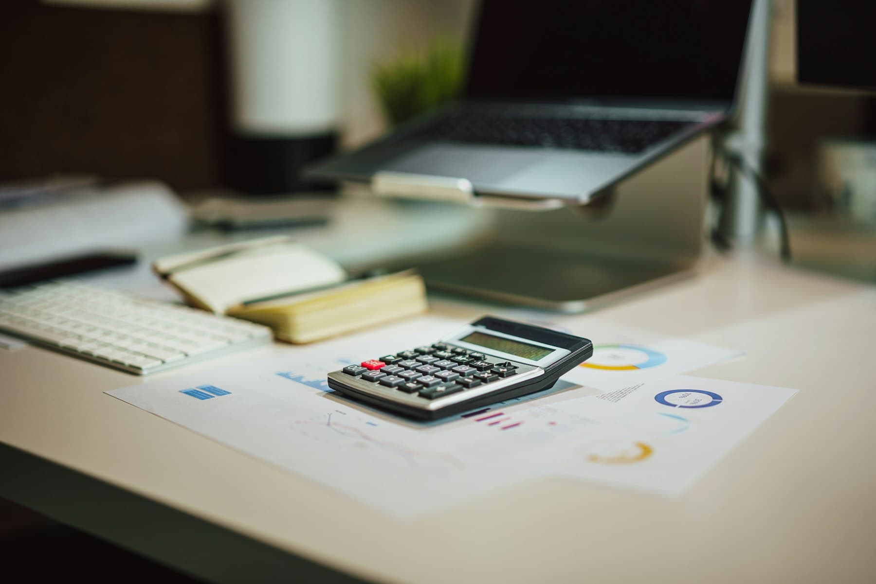 A calculator on printed reports on a work desk, with a laptop in the background